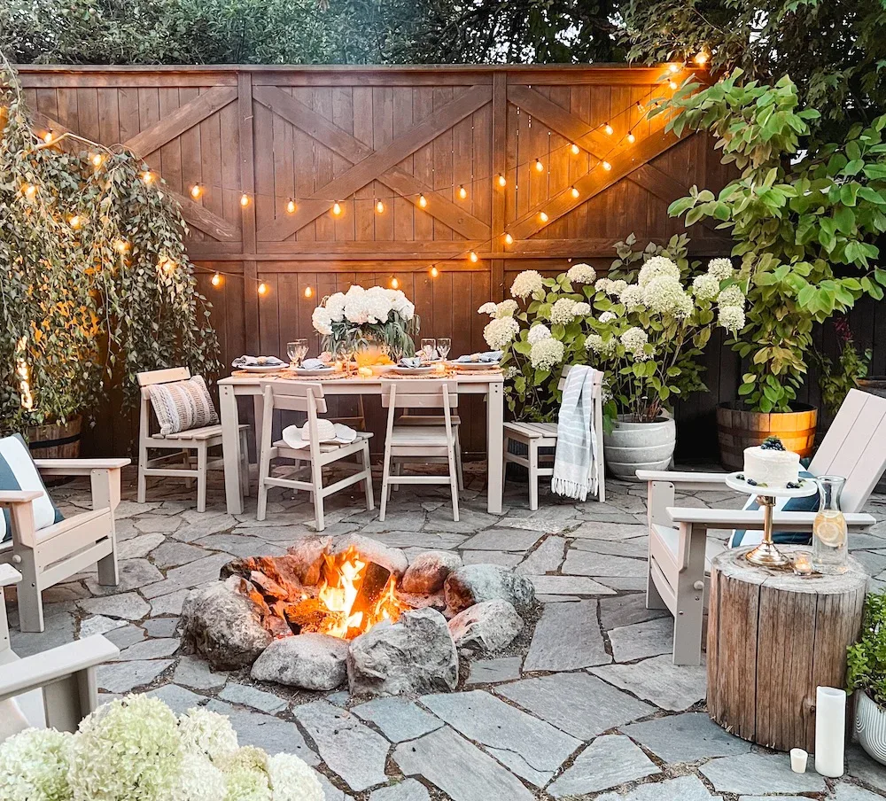 Entertaining outdoor patio dining with a table, wine, cake, florals,  white hydrangea florals and potted flower plants, stone pavement, fireplace, wood slap table and lighting