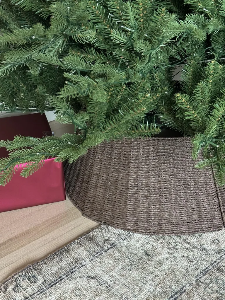 Beautiful Designer Christmas Decor Dupes You Can Buy or DIY – Natural Woven Christmas Tree Collar

A natural woven wicker collar surrounds the base of a green Christmas tree, concealing the stand with a simple, textured finish. Set on a neutral patterned rug, this understated accessory complements farmhouse, rustic, and minimalist holiday decorating styles.