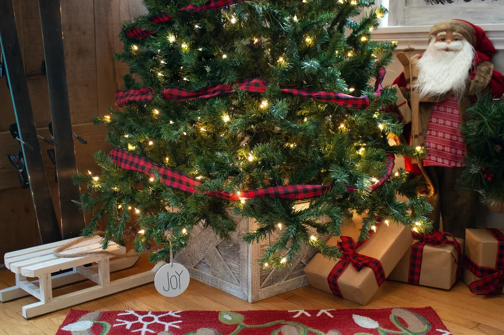 Beautiful Designer Christmas Decor Dupes You Can Buy or DIY – Traditional Christmas Tree with Plaid Ribbon

A lush Christmas tree glows with warm white lights and is decorated with red plaid ribbon garlands for a cozy, traditional holiday look. Wrapped gifts, a rustic wooden tree collar, a miniature white sled, and a Santa figure complete the festive display.