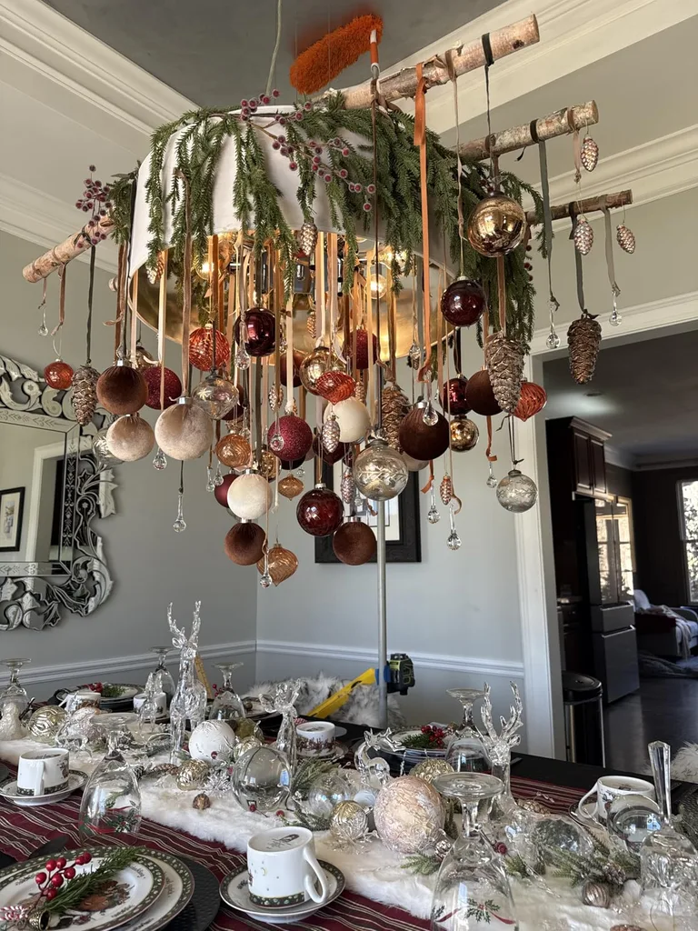 Beautiful Designer Christmas Decor Dupes You Can Buy or DIY – Christmas Ornament Chandelier

A decorative chandelier features an arrangement of hanging Christmas ornaments in metallic, copper, and neutral tones suspended above a dining table. Paired with festive greenery, elegant table settings, and a bright, airy interior, this creative display brings seasonal charm to the dining space.