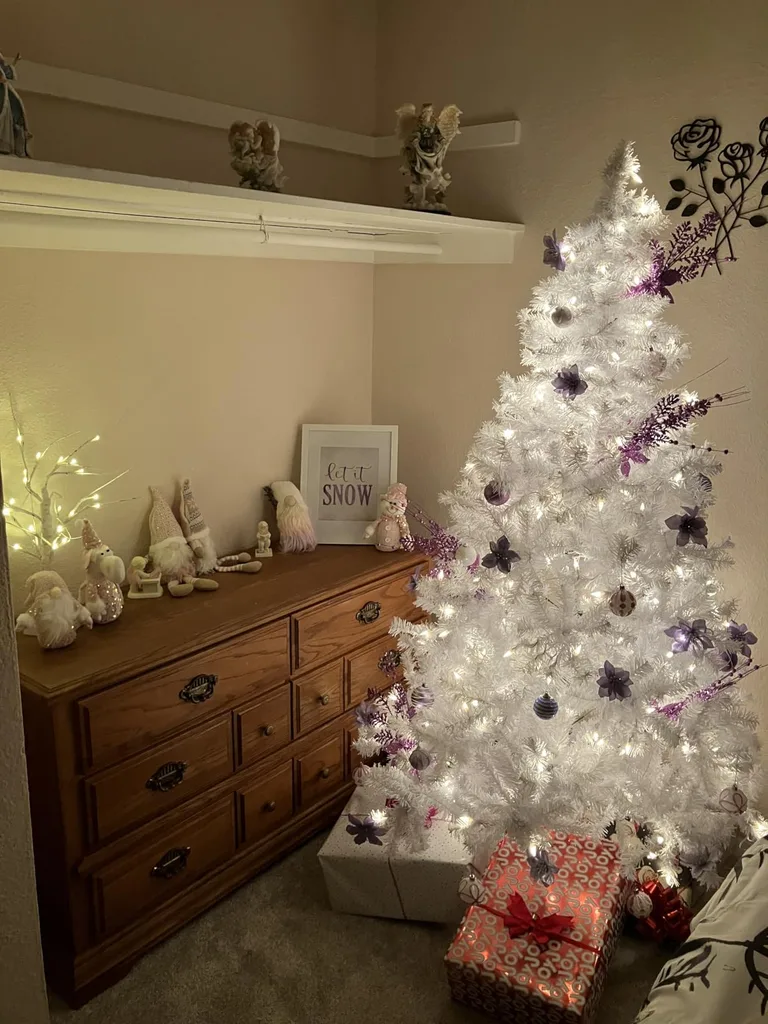 Beautiful Designer Christmas Decor Dupes You Can Buy or DIY – White Christmas Tree with Purple Ornaments

A white artificial Christmas tree glows with warm lights and is decorated with purple and silver ornaments for a soft, winter-inspired look. Positioned beside a dark wooden dresser with neutral seasonal decorations and wrapped presents underneath, the tree creates an elegant focal point in the room.