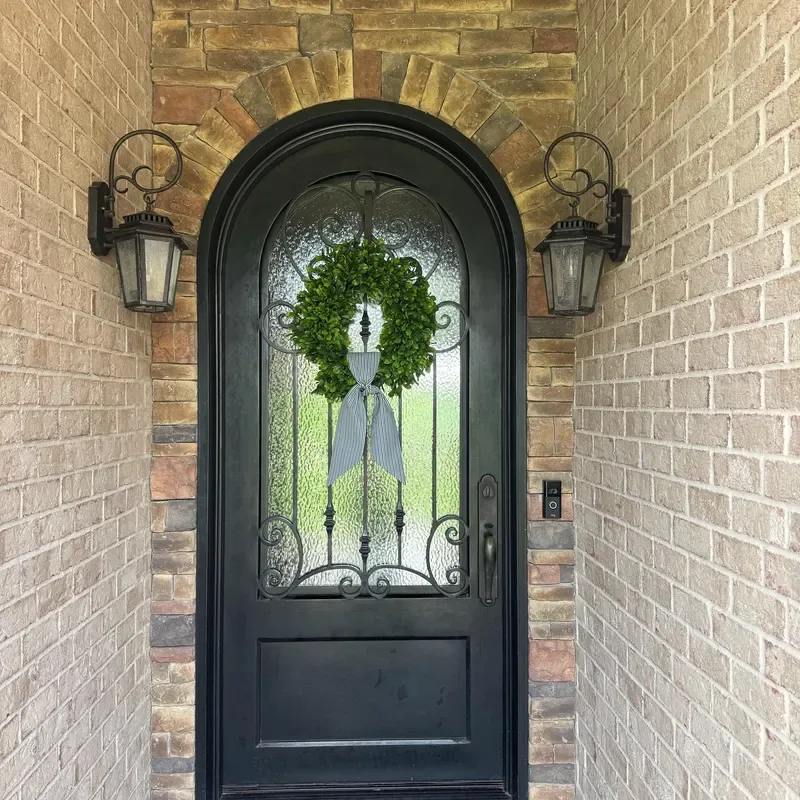 Easy Ways to Make Your Porch Feel Like Fall - A black arched front door with decorative glass panels and wrought-iron detailing, accented by a simple green wreath and warm brickwork for a timeless, welcoming entrance.