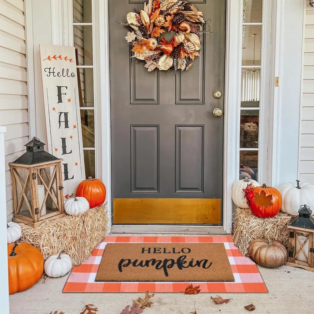 Easy Ways to Make Your Porch Feel Like Fall - Pumpkin-Themed Halloween Porch: A gray front door adorned with a colorful autumn wreath, surrounded by orange pumpkins, hay bales, rustic lanterns, and a layered orange-and-white doormat with a “Hello Pumpkin” design.