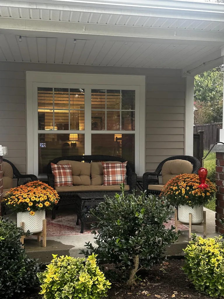 Easy Ways to Make Your Porch Feel Like Fall - Cozy Fall Front Porch Seating: A covered front porch with a neutral outdoor sofa, plaid throw pillows, and white planters filled with orange chrysanthemums, framed by neatly trimmed greenery and warm window lighting.