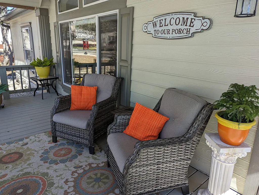 Easy Ways to Make Your Porch Feel Like Fall – A covered front porch with gray wicker seating, orange accent pillows, a colorful patterned rug, and potted greenery creates a comfortable outdoor sitting area with subtle fall touches.
