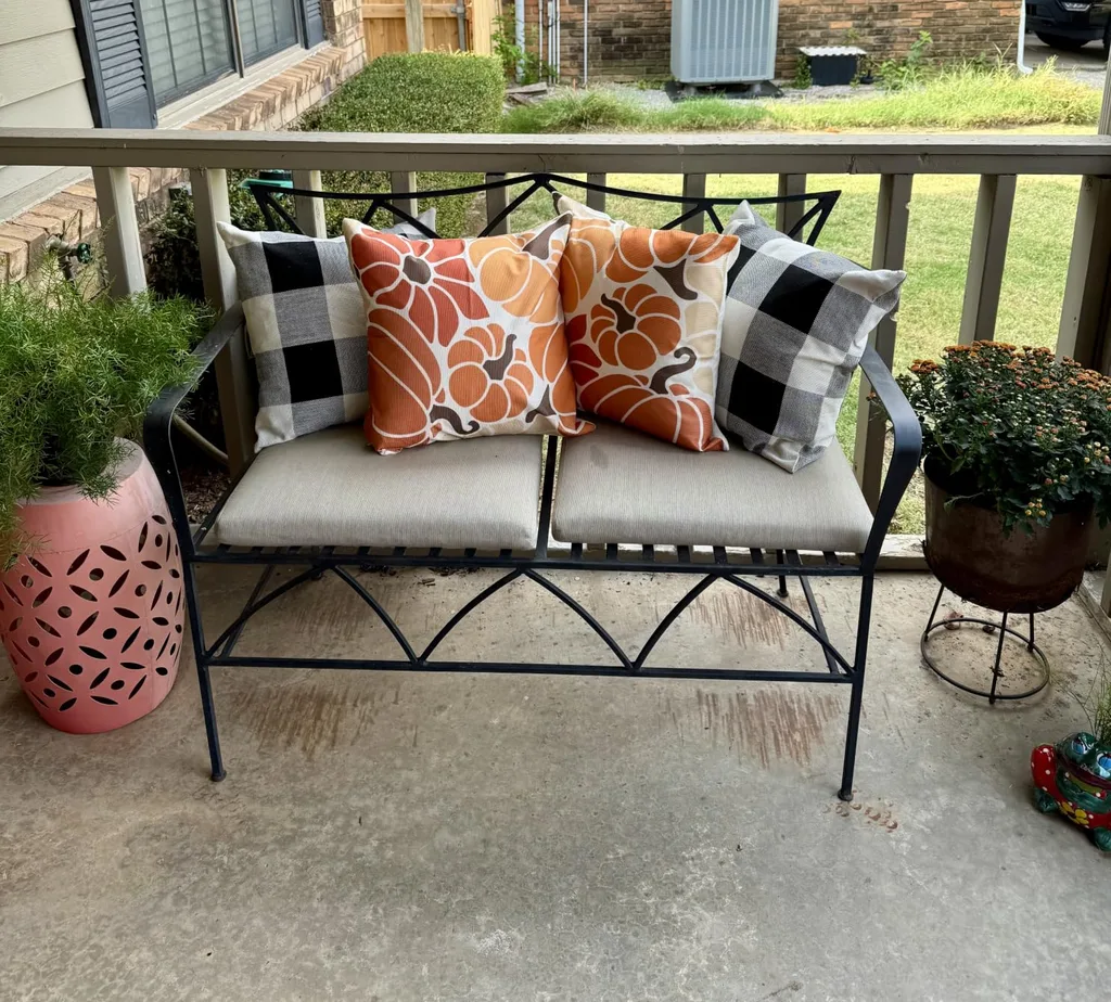 Easy Ways to Make Your Porch Feel Like Fall - Autumn Outdoor Bench Styling: A black metal patio bench with cream cushions, decorated with orange floral-patterned pillows and black-and-white buffalo plaid accents for a cozy fall seating area.