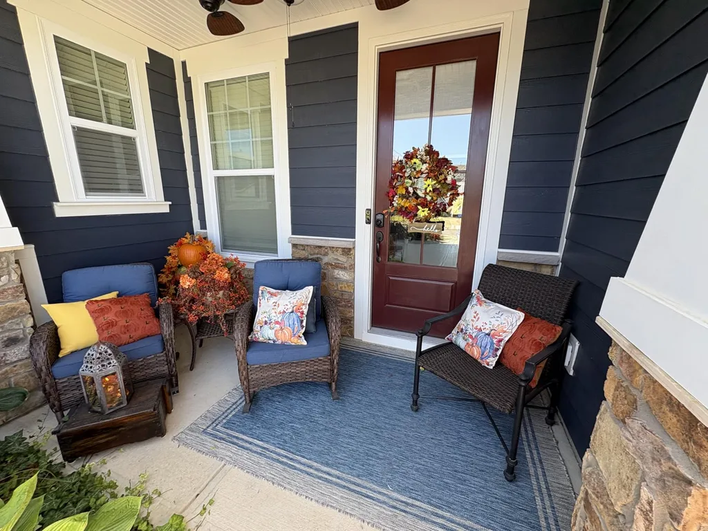 Easy Ways to Make Your Porch Feel Like Fall - Blue Porch with Seasonal Accents: A welcoming covered porch with navy siding, wicker chairs, colorful autumn throw pillows, a blue area rug, and orange fall foliage arranged beside a rich wood front door.