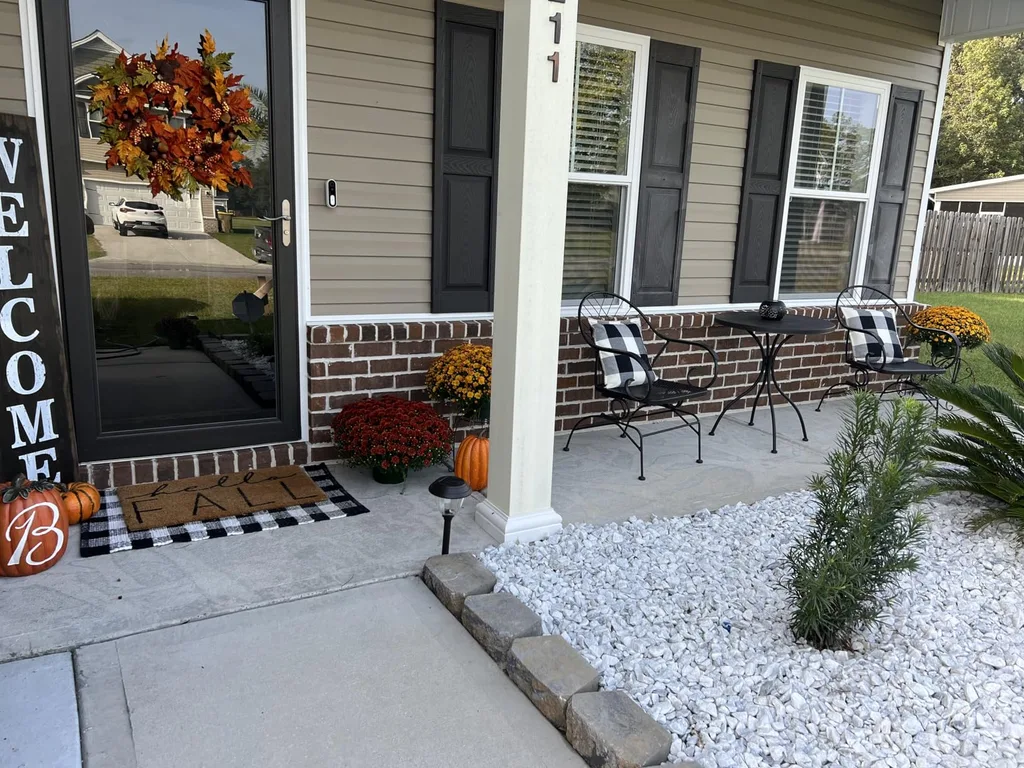 Easy Ways to Make Your Porch Feel Like Fall - Simple Fall Porch Decor: A modern home entrance featuring gray siding, white stone landscaping, a black metal bench with plaid cushions, and an autumn wreath accented by orange leaves near the front door.