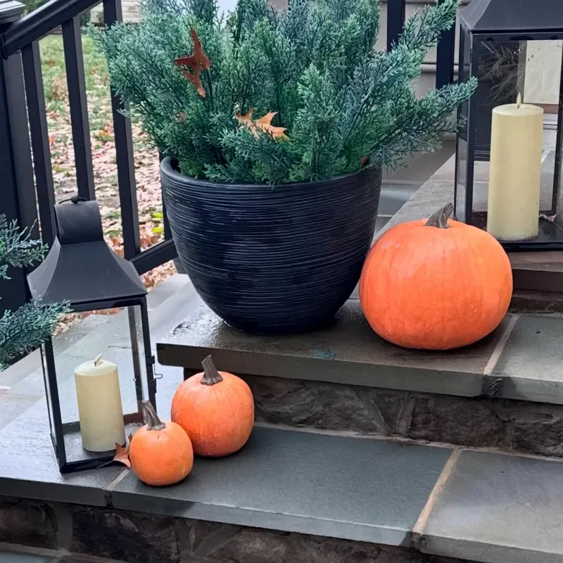 Easy Ways to Make Your Porch Feel Like Fall - Pumpkins and Potted Evergreens: A close-up of outdoor steps styled with a large black planter of evergreen foliage, orange pumpkins in varying sizes, and cream pillar candles for a simple seasonal display.