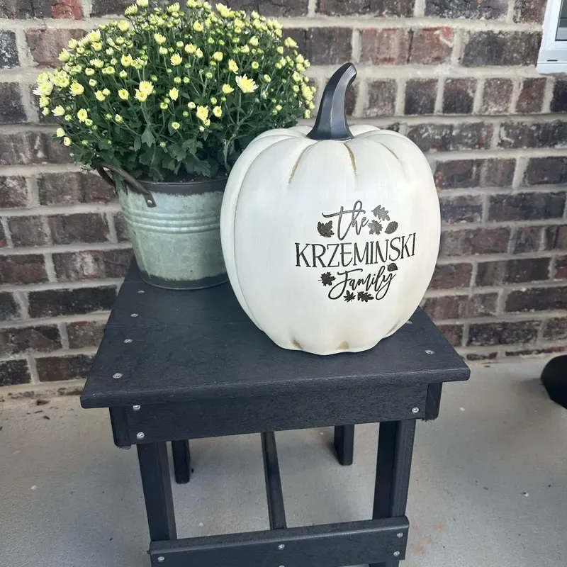 Easy Ways to Make Your Porch Feel Like Fall - White pumpkin personalized with a family name displayed on a black outdoor table beside a potted yellow mum against a brick wall.