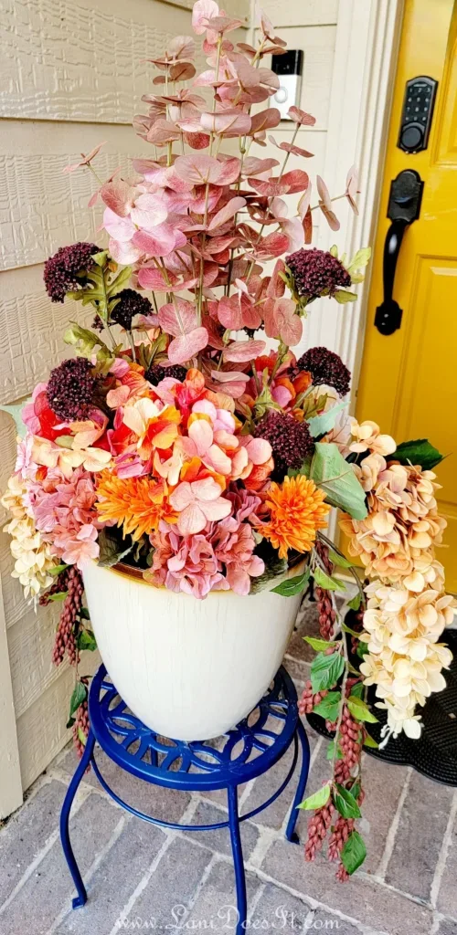 Fall in Love With This Gorgeous Fall-O-Ween Floral DIY - White ceramic planter overflowing with pink, orange, and red flowers, elevated on a decorative blue metal plant stand.