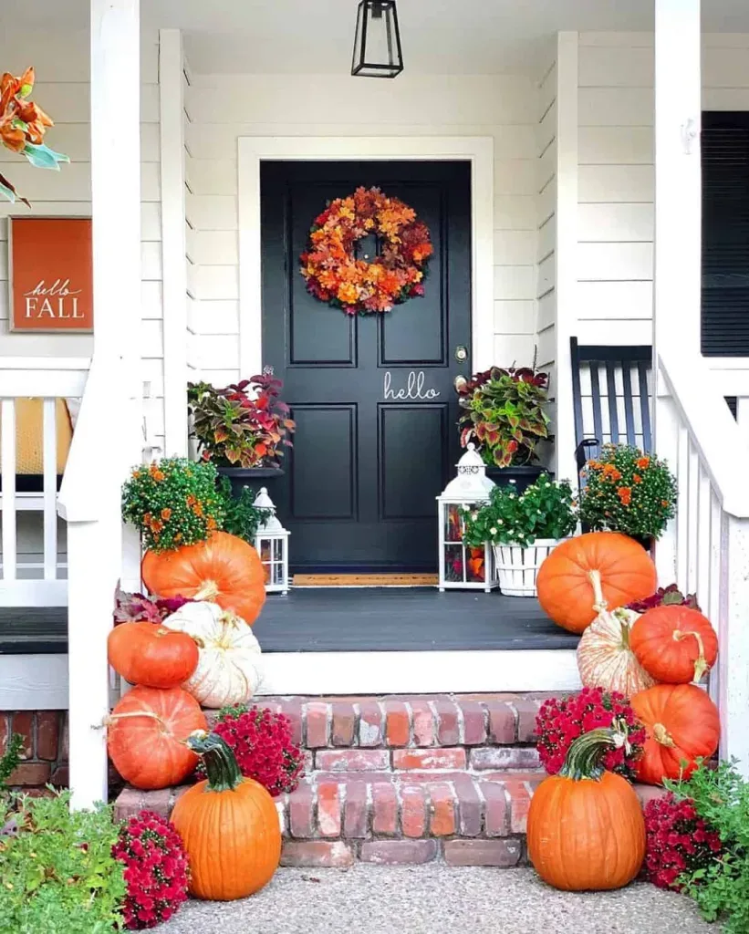 Easy Ways to Make Your Porch Feel Like Fall - White farmhouse porch with a black front door, autumn wreath, orange pumpkins lining brick steps, colorful potted flowers, and black rocking chairs.