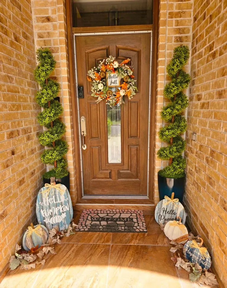 Easy Ways to Make Your Porch Feel Like Fall - Wooden front door decorated with an autumn wreath, flanked by potted greenery, rustic pumpkins, and seasonal welcome signs.