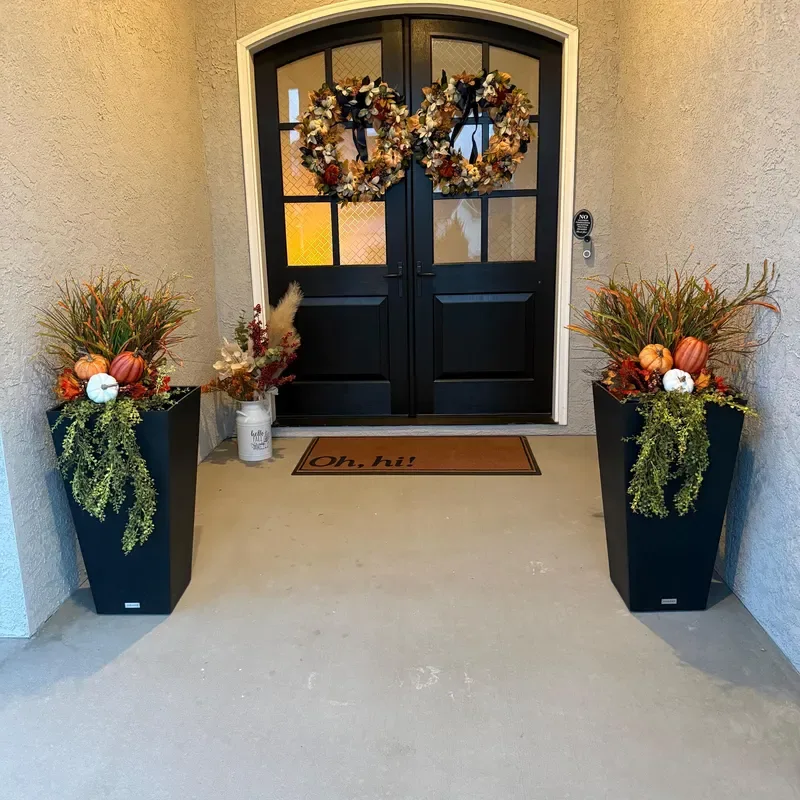 Easy Ways to Make Your Porch Feel Like Fall - A black double front door with glass panels and autumn wreaths creates a striking focal point in this covered entryway. Tall black planters filled with ornamental grasses, cascading greenery, orange foliage, and white pumpkins frame the entrance, adding seasonal color and texture. A simple fall doormat completes the polished, welcoming look.