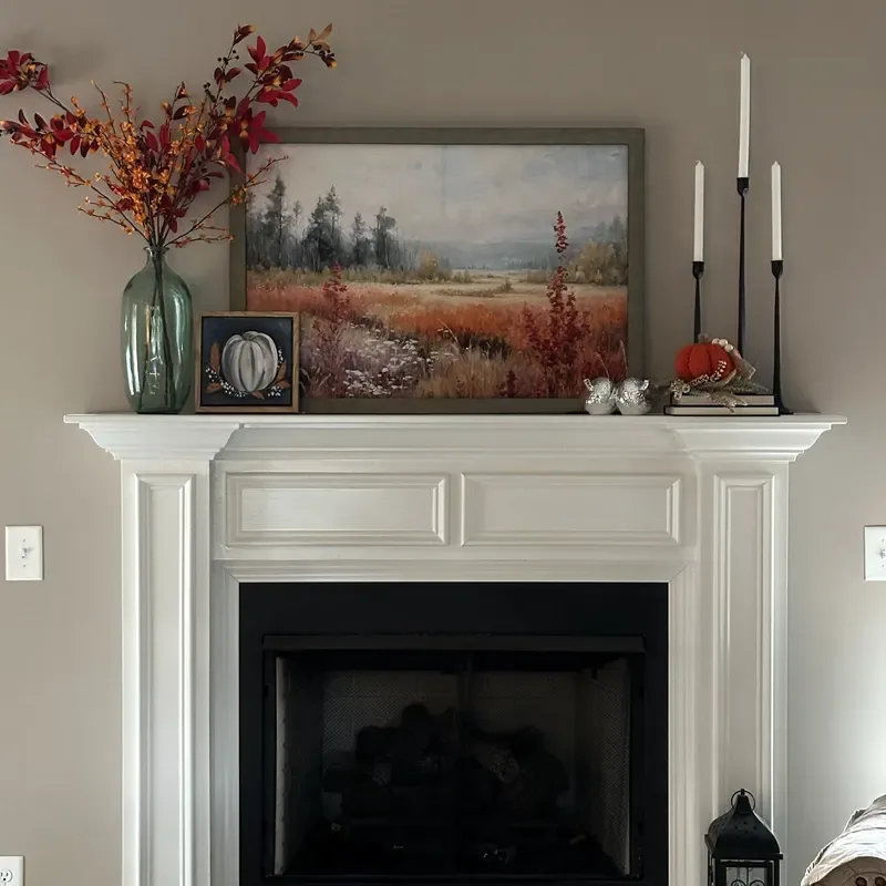 Refresh Your Living Room for Fall in 7 Easy Ways – A white fireplace mantel decorated with autumn landscape artwork, a green glass vase filled with fall branches, white candles, and small pumpkins adds warm seasonal touches to a neutral living room.