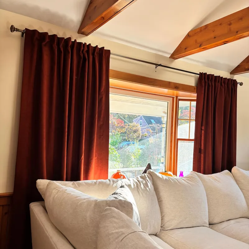 Refresh Your Living Room for Fall in 7 Easy Ways – Rich burgundy curtains frame a large living room window, adding warmth and contrast to the cream sofa, exposed wooden ceiling beams, and bright natural light.