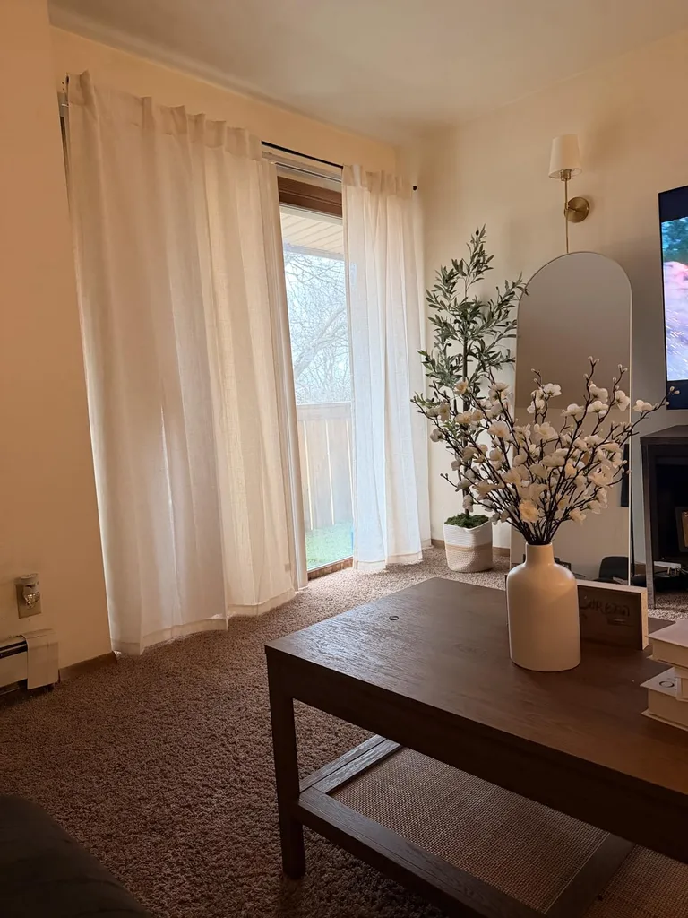 Refresh Your Living Room for Fall in 7 Easy Ways – Sheer cream curtains soften the natural light in a neutral living room, while a wooden coffee table, decorative branches, and a tall indoor plant add texture and a relaxed seasonal feel.