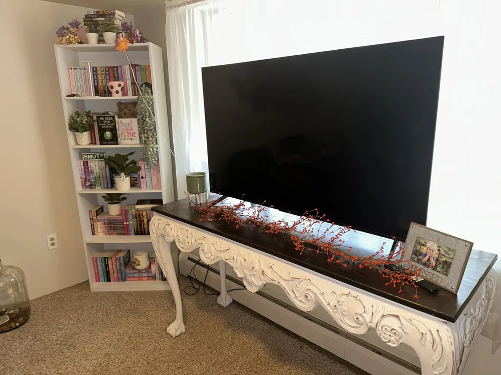 Refresh Your Living Room for Fall in 7 Easy Ways – A large television sits on an ornate white console table beside a bookshelf filled with colorful decor and books, offering a simple living room setup that can be refreshed with autumn accents.