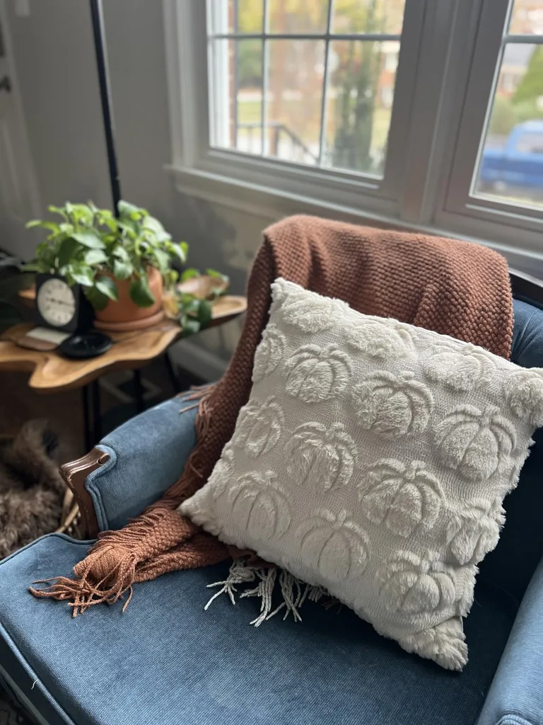 Refresh Your Living Room for Fall in 7 Easy Ways – A cozy accent chair beside a sunlit window features a textured cream pillow and a warm rust-colored throw, creating a comfortable reading nook with natural light and subtle fall colors.