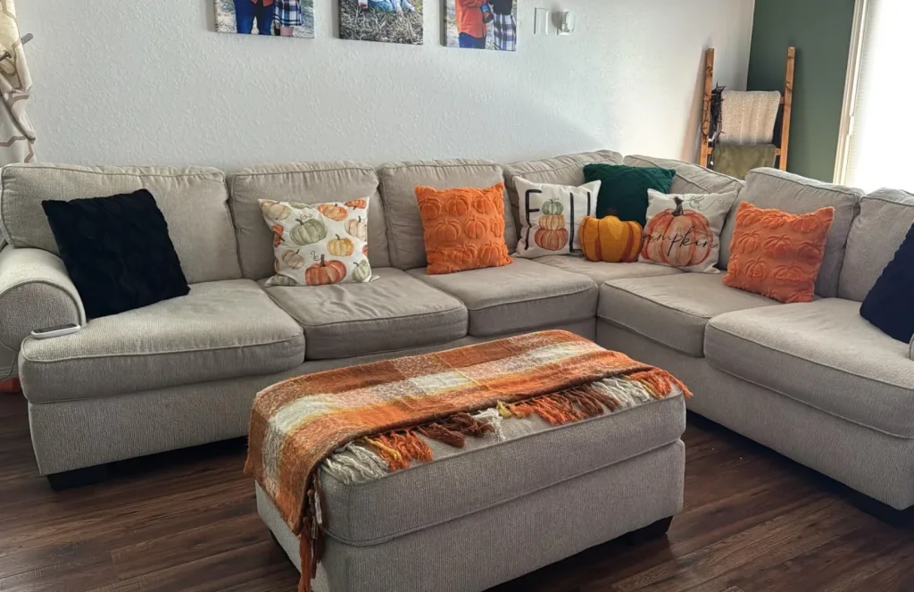 Refresh Your Living Room for Fall in 7 Easy Ways – A gray sectional sofa decorated with orange, rust, cream, and dark accent pillows pairs with a matching ottoman topped with a warm-toned throw, bringing cozy autumn colors into a modern living room.