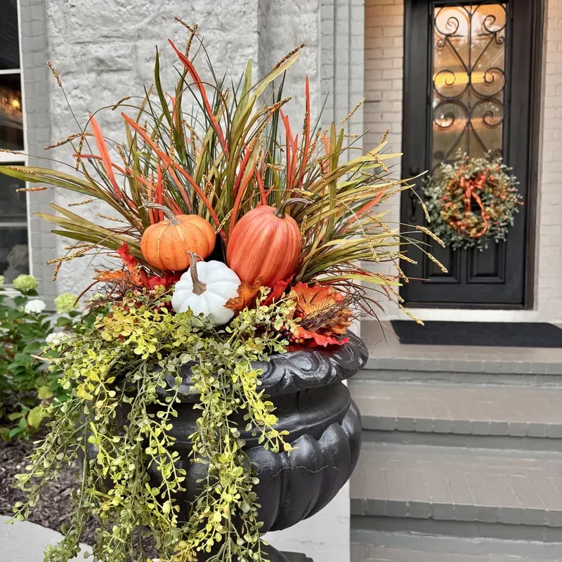 Easy Ways to Make Your Porch Feel Like Fall - arge black planter filled with ornamental grasses, orange and white pumpkins, and cascading greenery beside a home's front entrance.