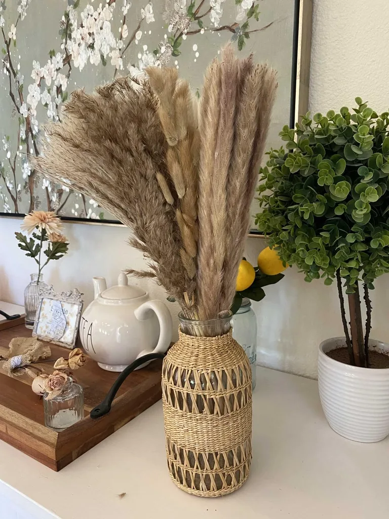 Refresh Your Living Room for Fall in 7 Easy Ways – A woven vase filled with tall dried pampas grass sits on a wooden console table alongside white ceramic decor, a leafy potted plant, and a floral wall accent for a warm, natural look.