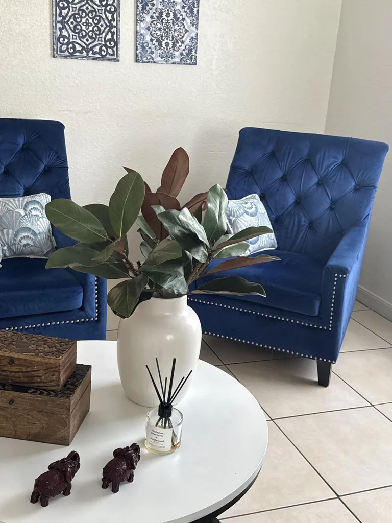 Refresh Your Living Room for Fall in 7 Easy Ways – A white ceramic vase with broad green and burgundy leaves sits on a round white coffee table between blue upholstered armchairs, creating a fresh botanical accent in the living room.