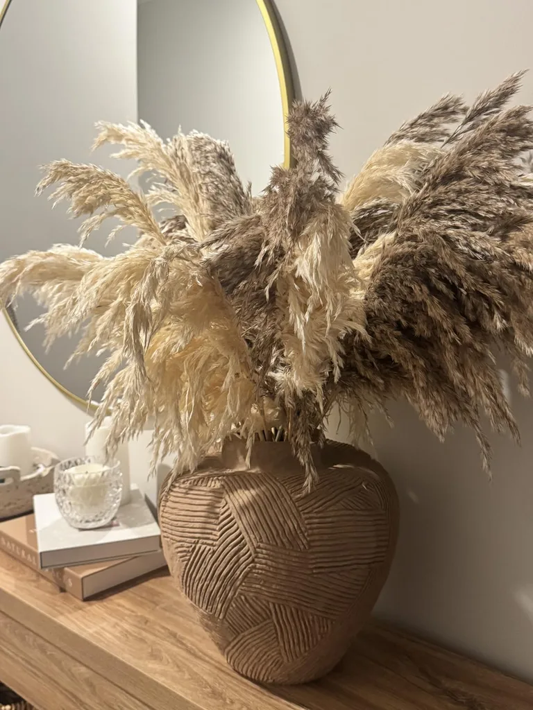 Refresh Your Living Room for Fall in 7 Easy Ways – Tall dried pampas grass arranged in a textured brown vase adds softness and natural warmth to a wooden side table beside a round mirror and neutral decorative accessories.