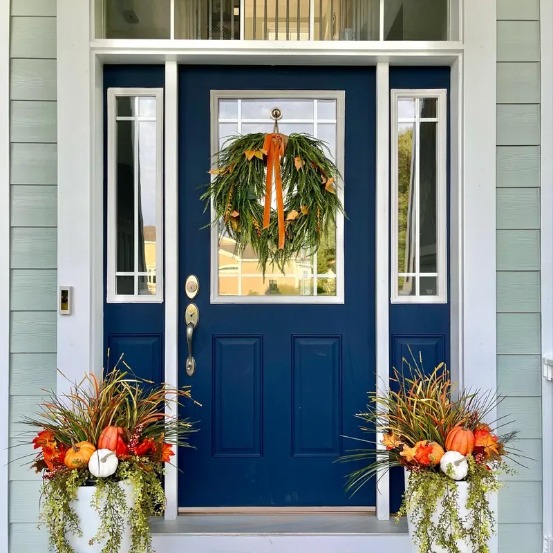 Easy Ways to Make Your Porch Feel Like Fall - Navy blue front door decorated with a lush autumn wreath and orange ribbon, flanked by matching planters filled with ornamental grasses, orange flowers, and white pumpkins.