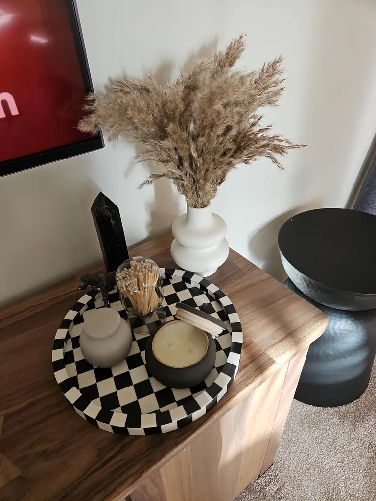 Refresh Your Living Room for Fall in 7 Easy Ways – A wooden side table styled with a white vase of dried pampas grass, neutral decorative objects, and a black-and-white checkered tray creates a cozy arrangement with natural textures.
