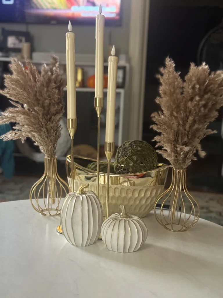 Refresh Your Living Room for Fall in 7 Easy Ways – Dried pampas grass, cream pillar candles in gold holders, and white decorative pumpkins create an elegant fall display on a light-colored coffee table.