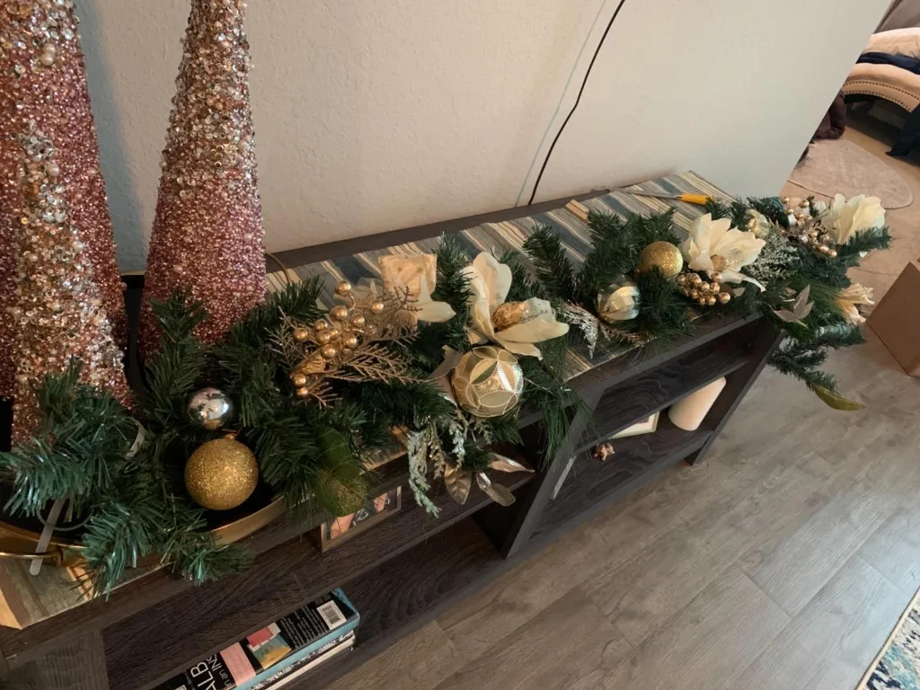Beautiful Designer Christmas Decor Dupes You Can Buy or DIY – A dark wooden console table is styled with lush evergreen garland, gold ornaments, cream accents, and sparkling decorative trees for an elegant Christmas display.