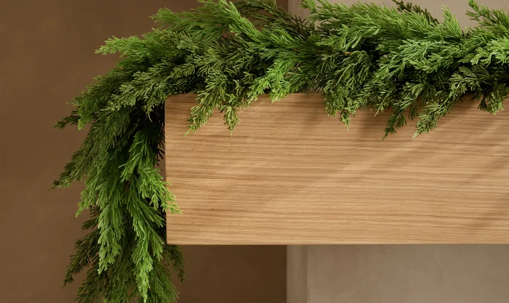 Beautiful Designer Christmas Decor Dupes You Can Buy or DIY – A thick evergreen garland drapes across a light wooden mantel, offering a simple, natural Christmas decoration that can inspire an affordable DIY alternative to designer greenery.