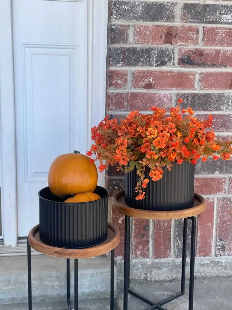 Easy Ways to Make Your Porch Feel Like Fall - A black ribbed planter filled with orange autumn flowers sits on a raised wooden stand beside a smaller matching planter holding a pumpkin, creating a simple seasonal display against a brick exterior.