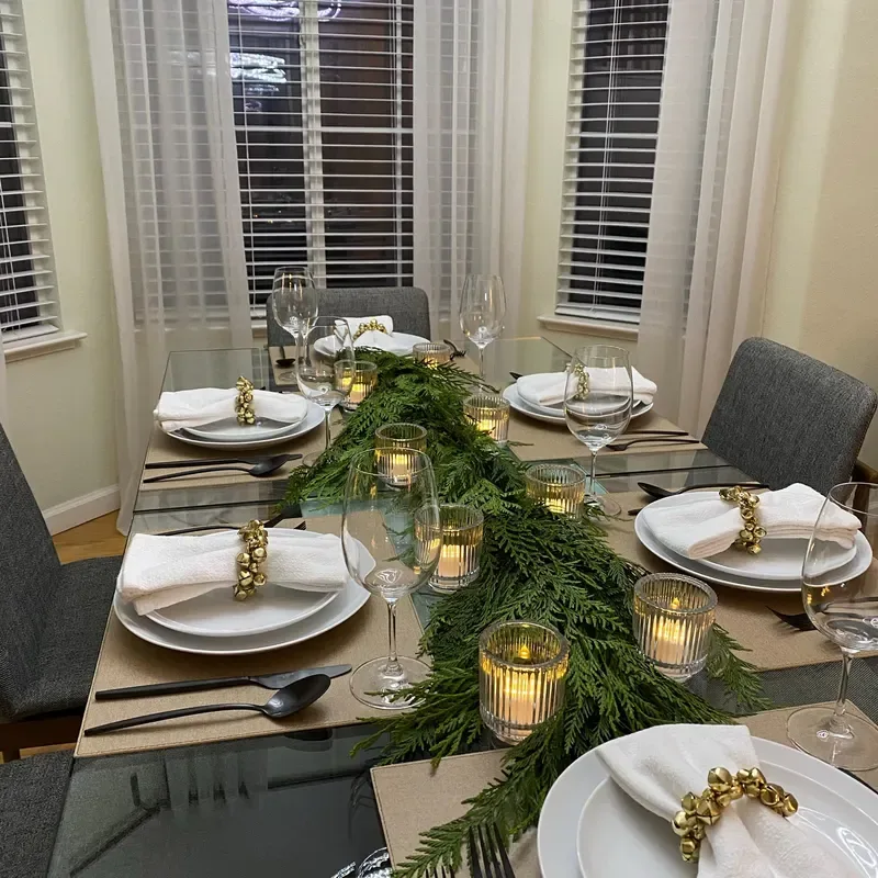 Beautiful Designer Christmas Decor Dupes You Can Buy or DIY – A dining table near a window is styled with white plates, gold flatware, green foliage, and glowing candles in glass holders, creating an elegant Christmas tablescape with natural greenery and warm metallic accents.