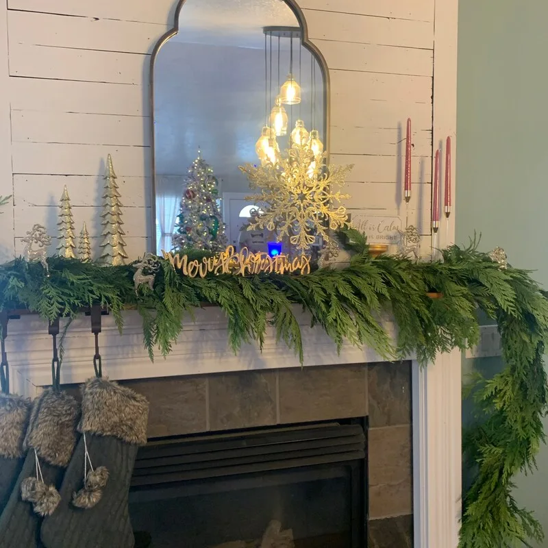 Beautiful Designer Christmas Decor Dupes You Can Buy or DIY – A white shiplap fireplace mantel is decorated with cascading evergreen garland, gold decorative trees, a large gold snowflake ornament, and a gold “Merry Christmas” sign. Red taper candles and gray fur-trimmed stockings complete the festive display, creating an elegant Christmas look with rustic farmhouse charm.