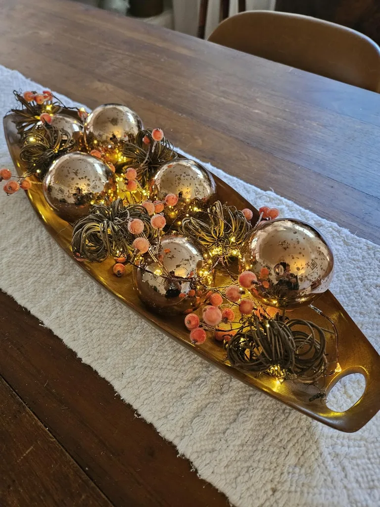 Beautiful Designer Christmas Decor Dupes You Can Buy or DIY – Festive Christmas Table Centerpiece

A long decorative tray sits on a wooden table, filled with gold Christmas ornaments, pinecones, dried citrus slices, and seasonal greenery. This natural arrangement brings warm, rustic charm to a holiday tablescape or dining room centerpiece.